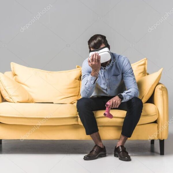 upset man lost in video game with joystick and virtual reality headset on yellow sofa on grey