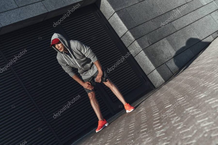 young man in sports clothing resting after training in street at stone wall