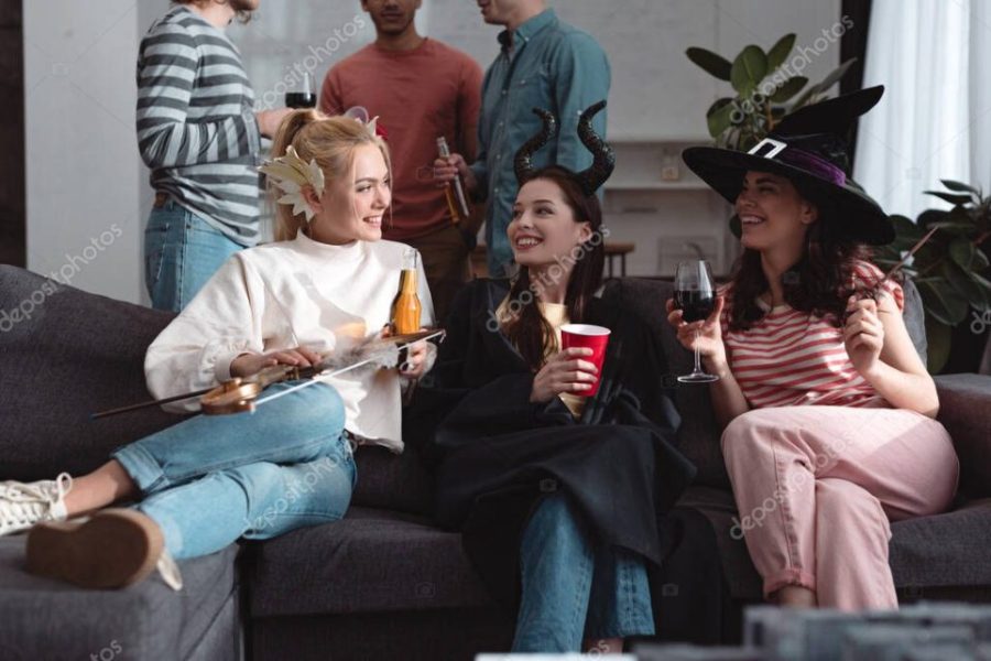 cropped view of men standing near cheerful girls in fairy costumes drinking beverages on sofa