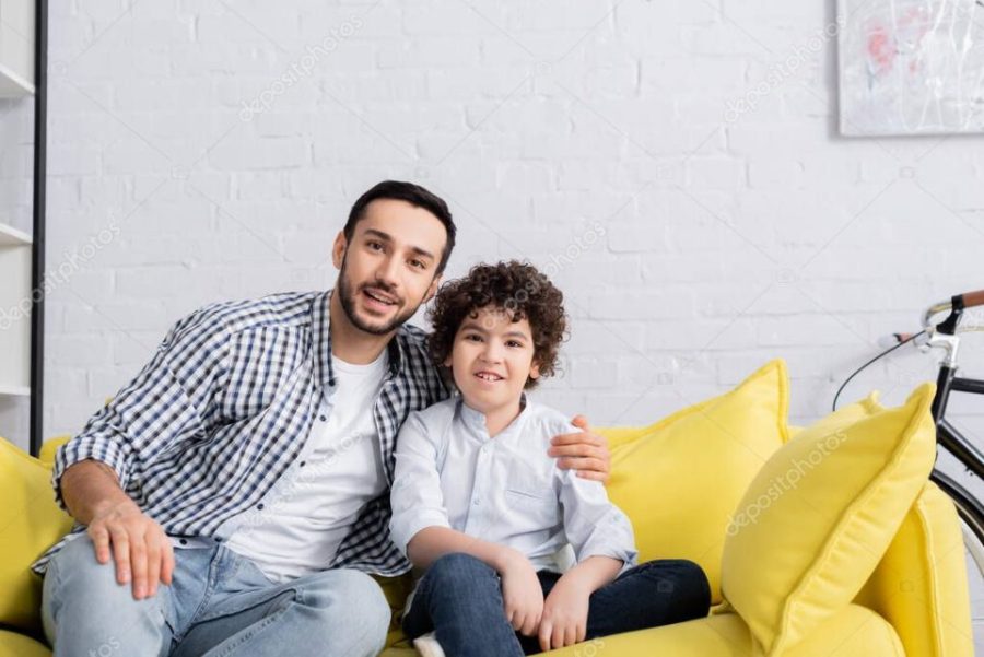 happy muslim man embracing son while sitting on sofa and looking at camera