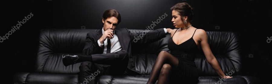man in suit sitting on leather sofa with glass of whiskey near woman in slip dress on black, banner