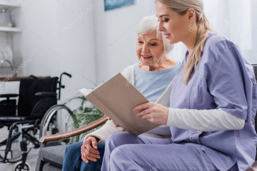 young social worker reading book to senior woman while sitting on sofa