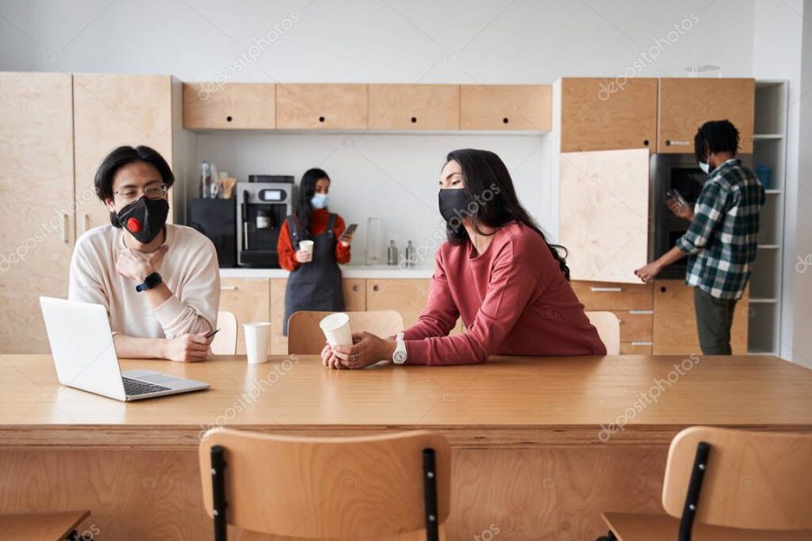 Two multiracial students wearing protective masks sitting at the table at the college kitchen