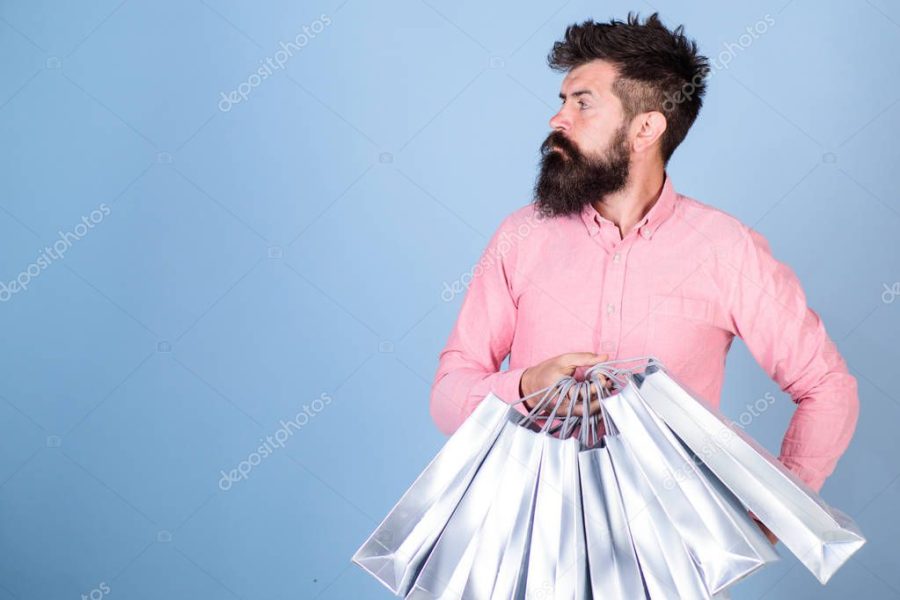 Man in pink shirt with stylish beard isolated on blue background. Hipster with trendy beard and mustache waiting for someone. Bearded man carrying silver shopping bags in hands, fashion concept