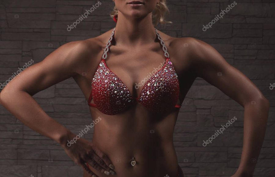 Sexy blond bodybuilder woman in red bikini with crystals over stone wall background shows muscles, closeup chest stomuch and arms