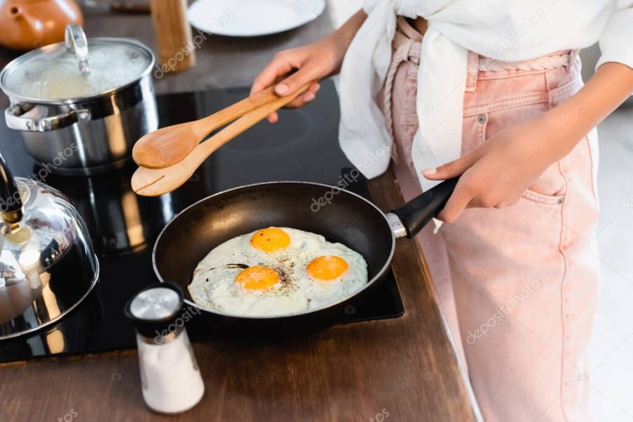 cropped view of woman holding kitchen tongs near eggs on frying pan