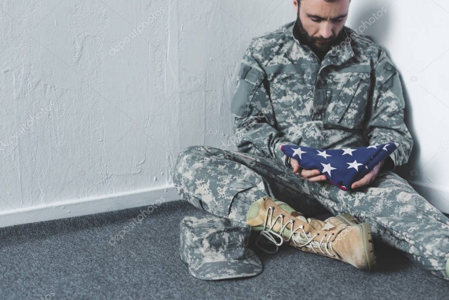 sad bearded man in military uniform sitting on floor in corner and holding usa national flag