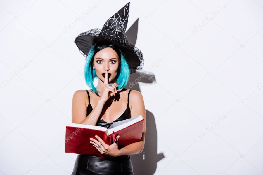 sexy girl in black witch Halloween costume with blue hair holding book and showing shh gesture on white background