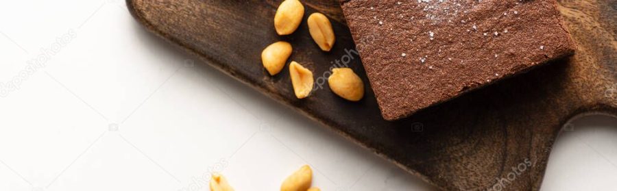top view of delicious brownie piece on wooden cutting board with nuts on white background, panoramic shot