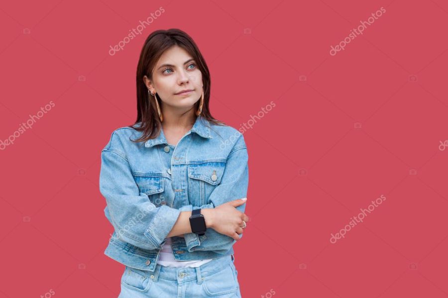 Portrait of thoughtful beautiful brunette young woman with makeup in denim casual style standing with crossed hands and looking away on red background.