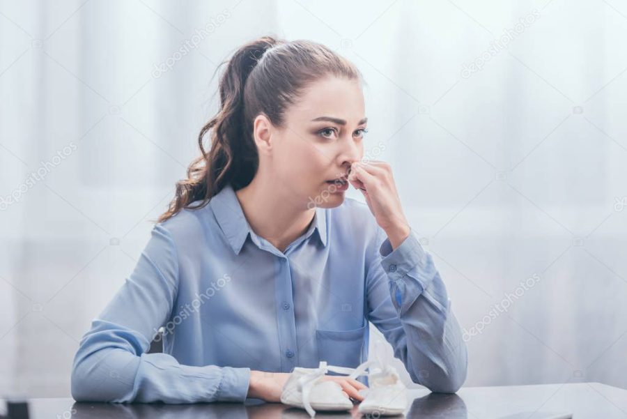 sad woman in blue blouse sitting at wooden table with baby shoes, crying and looking into distance at home, grieving disorder concept
