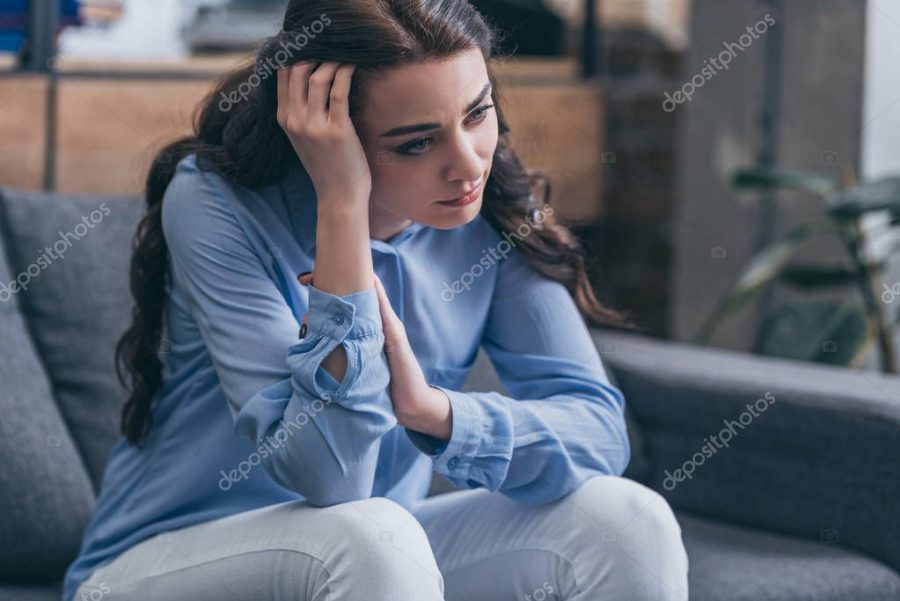 sad woman in blue blouse sitting on grey couch and looking into distance at home, grieving disorder concept