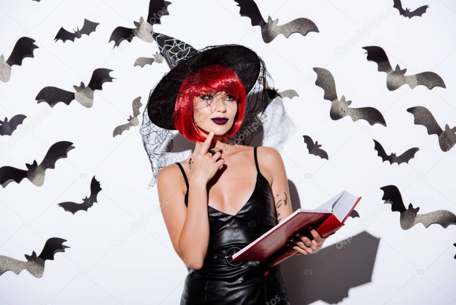 thoughtful girl in black witch Halloween costume with red hair holding book near white wall with decorative bats