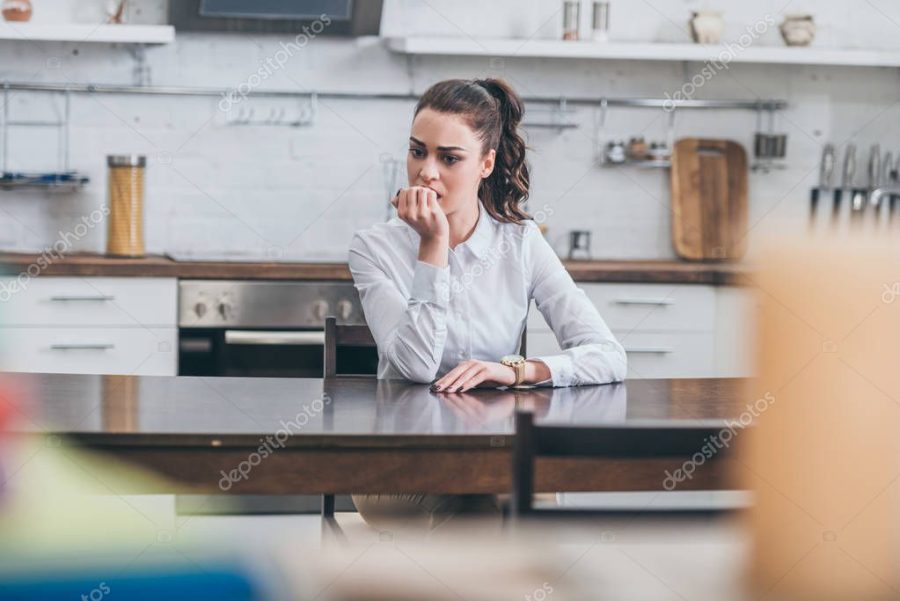 upset woman in white blouse sitting ar table and bitting nails in kitchen, grieving disorder concept