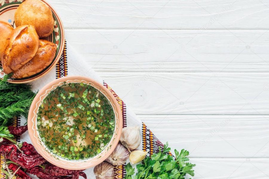 top view of traditional fish soup with green onion, embroidered towel, ingredients and mini pies on white wooden background