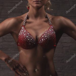 Sexy blond bodybuilder woman in red bikini with crystals over stone wall background shows muscles, closeup chest stomuch and arms