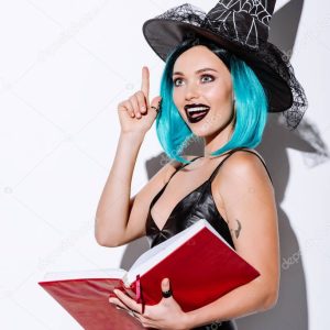 excited sexy girl in black witch Halloween costume with blue hair holding book and showing idea gesture on white background