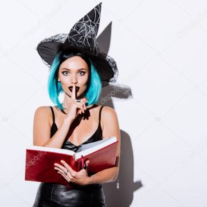 sexy girl in black witch Halloween costume with blue hair holding book and showing shh gesture on white background