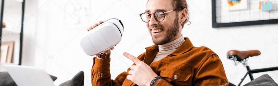 Panoramic shot of smiling 3d artist pointing on vr headset while having video call on laptop