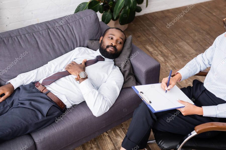 high angle view of african american man in formal wear lying on sofa near psychologist writing on clipboard