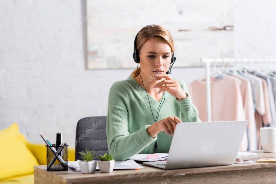 Blonde freelancer in headset having video chat on laptop on blurred foreground