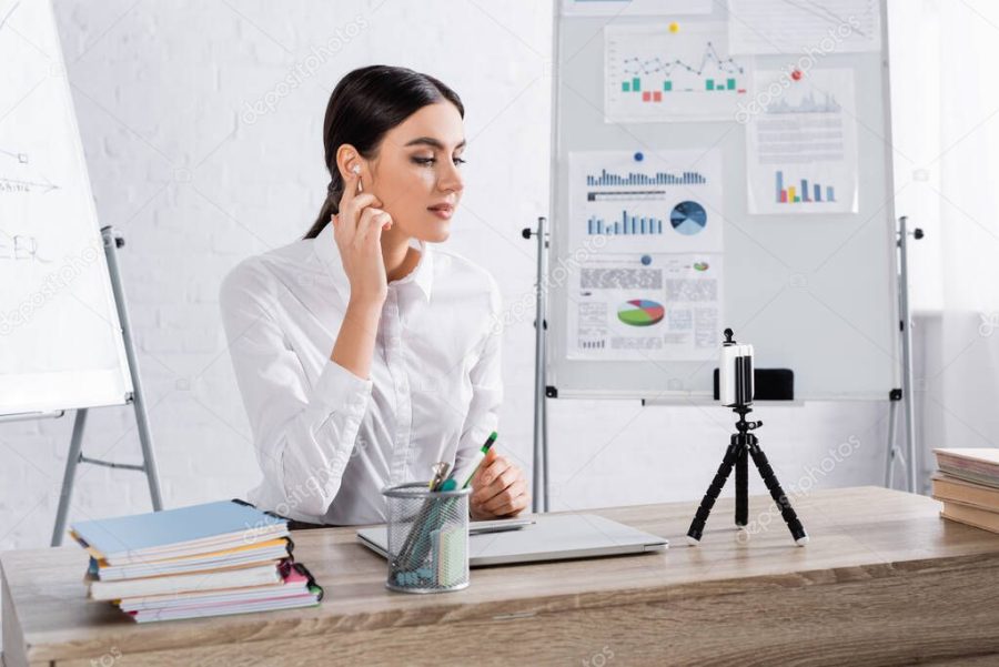 Businesswoman in earphone looking at smartphone during video call in office