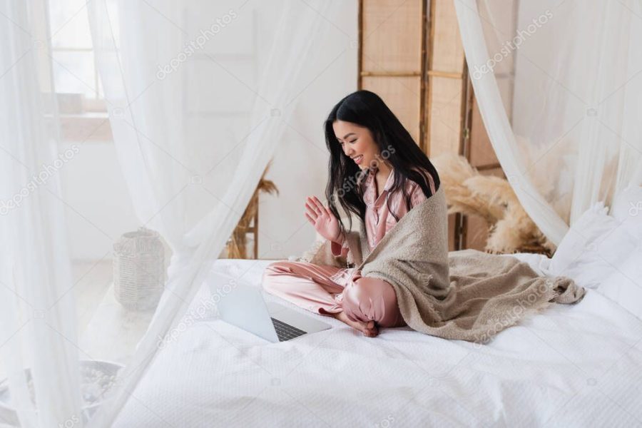 cheerful young asian woman sitting on bed and waving hand while having video call with laptop in bedroom