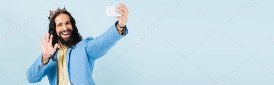 happy hispanic man in jacket and crown holding smartphone and having video call isolated on blue, banner