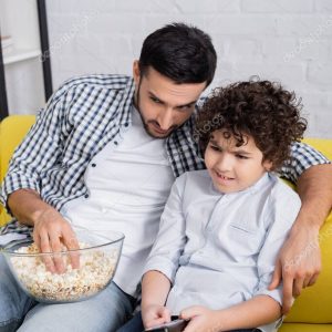 scared arabian boy holding tv remote controller while watching movie with father at home