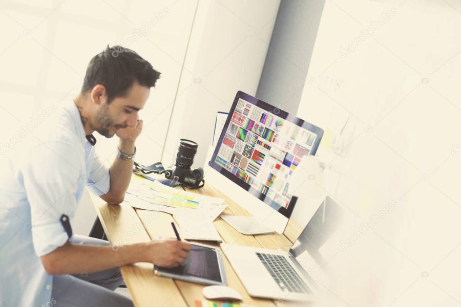 Portrait of young designer sitting at graphic studio in front of laptop and computer while working online.