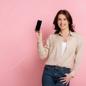 Beautiful brunette woman smiling at camera while holding smartphone on pink background